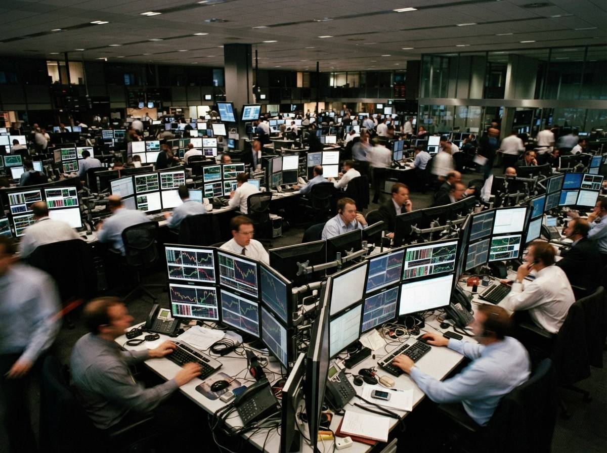 Traders sit at multi monitor desks on a dimly lit bank trading floor.